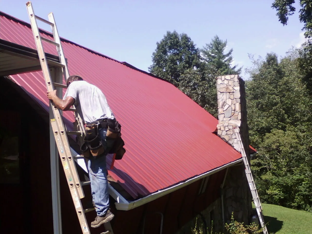 Roofer on a ladder beside a red metal roof and stone chimney — Soffit & Fascia Repair by Roofing Brothers in State Center